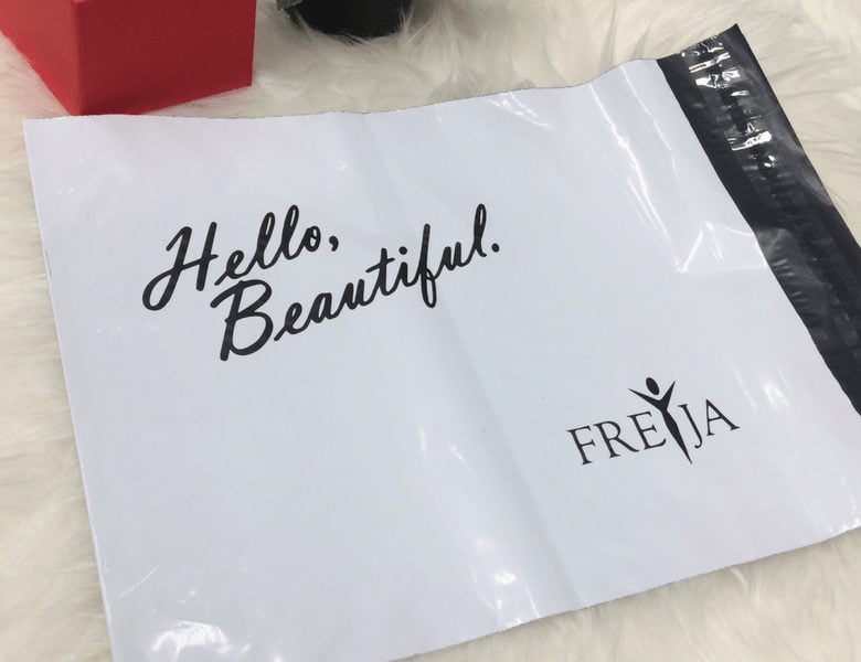 A white poly mailer bag with the text Hello, Beautiful. and the brand name FREJA printed on it, resting on a white faux fur surface.