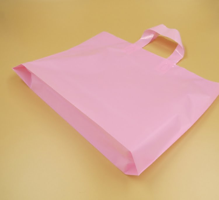 A flat, empty pink plastic shopping bag with die-cut handles lying on a tan surface.