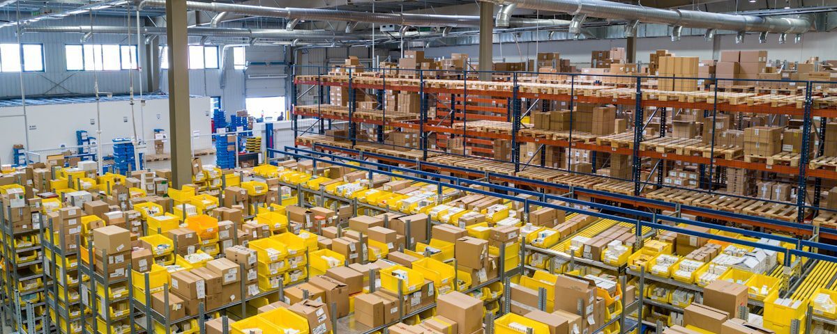 High-angle view of a large warehouse interior filled with rows of metal shelving units containing cardboard boxes and yellow plastic bins.