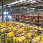 High-angle view of a large warehouse interior filled with rows of metal shelving units containing cardboard boxes and yellow plastic bins.