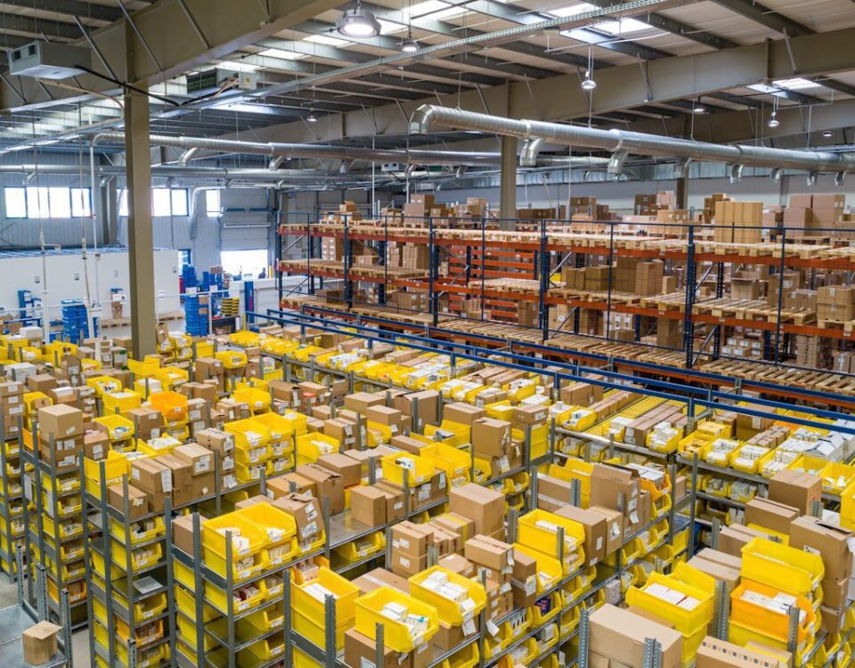 High-angle view of a large warehouse interior filled with rows of metal shelving units containing cardboard boxes and yellow plastic bins.