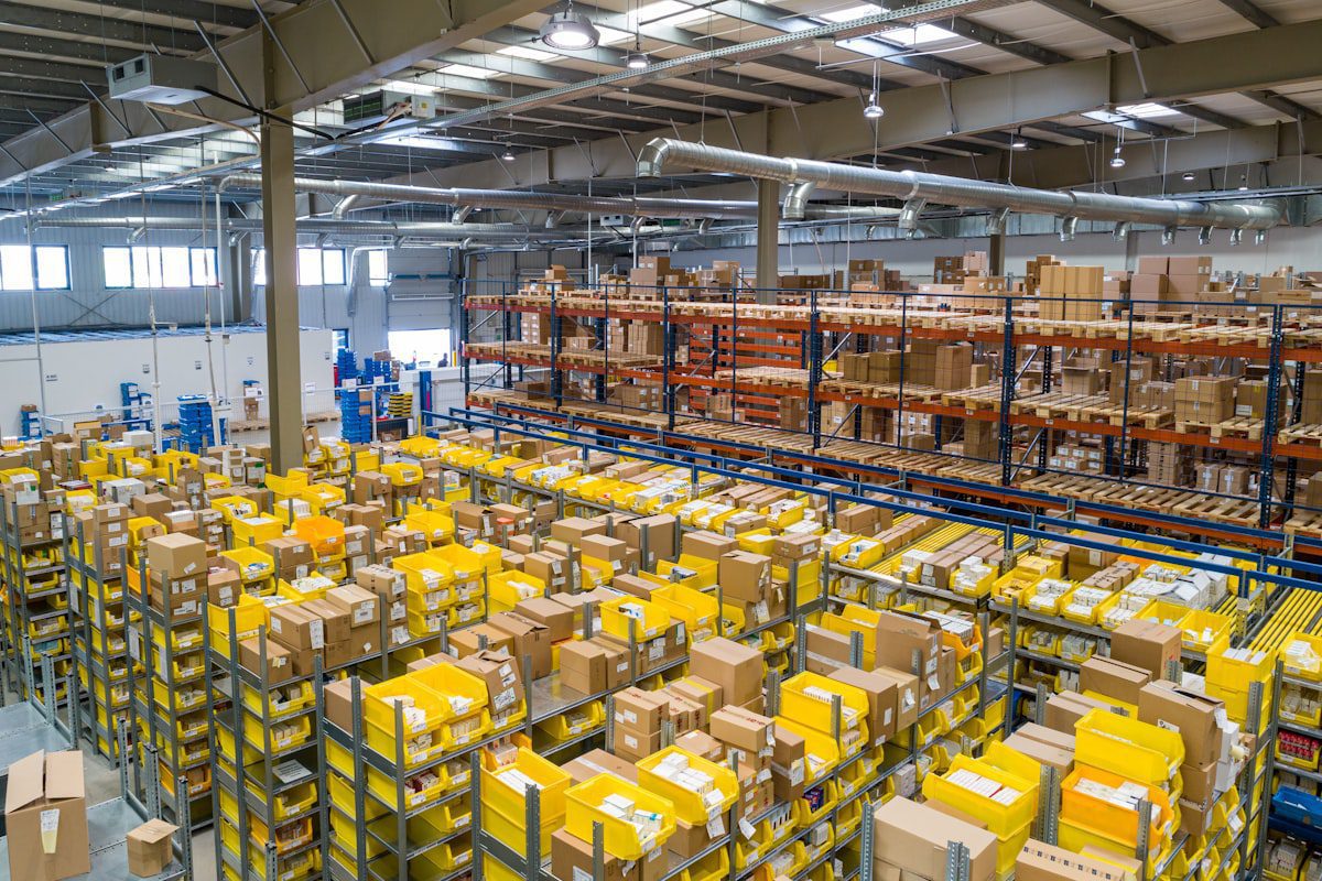High-angle view of a large warehouse interior filled with rows of metal shelving units containing cardboard boxes and yellow plastic bins.