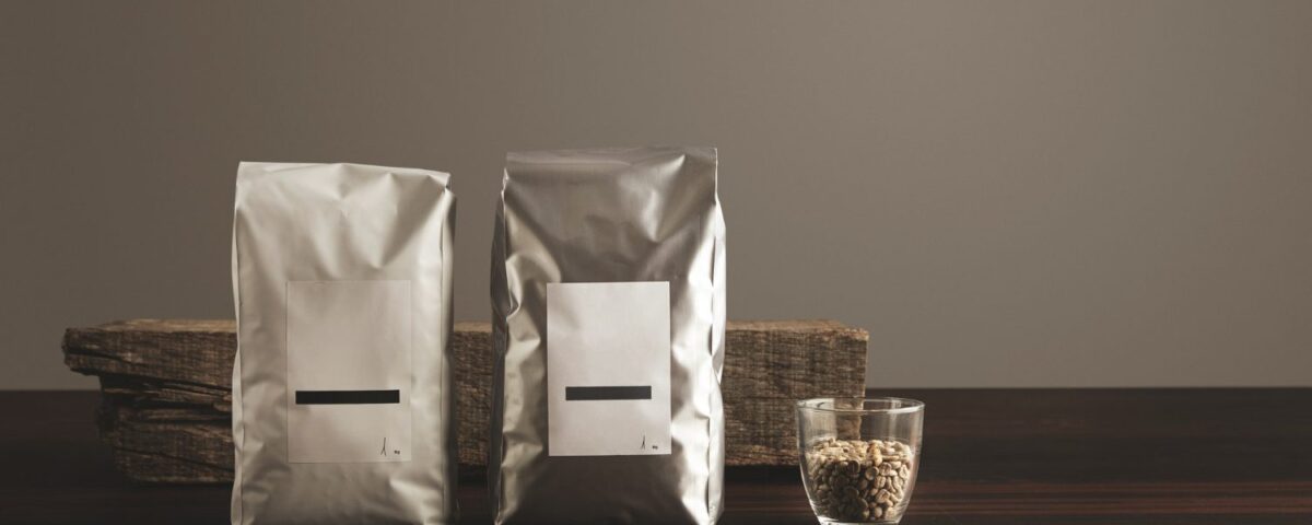 Two silver-colored pouches with blank white labels standing on a dark wooden surface next to a small glass filled with light-colored grains, with a piece of weathered wood in the background.