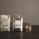 Two silver-colored pouches with blank white labels standing on a dark wooden surface next to a small glass filled with light-colored grains, with a piece of weathered wood in the background.