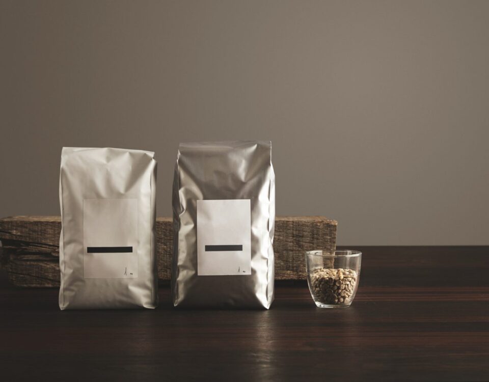 Two silver-colored pouches with blank white labels standing on a dark wooden surface next to a small glass filled with light-colored grains, with a piece of weathered wood in the background.