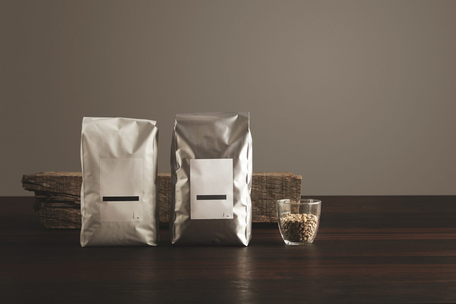 Two silver-colored pouches with blank white labels standing on a dark wooden surface next to a small glass filled with light-colored grains, with a piece of weathered wood in the background.