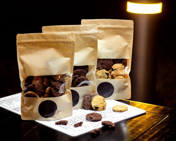 Three standing kraft paper pouches with clear windows filled with chocolate chip cookies, sitting on a white paper surface with loose cookies scattered in front.