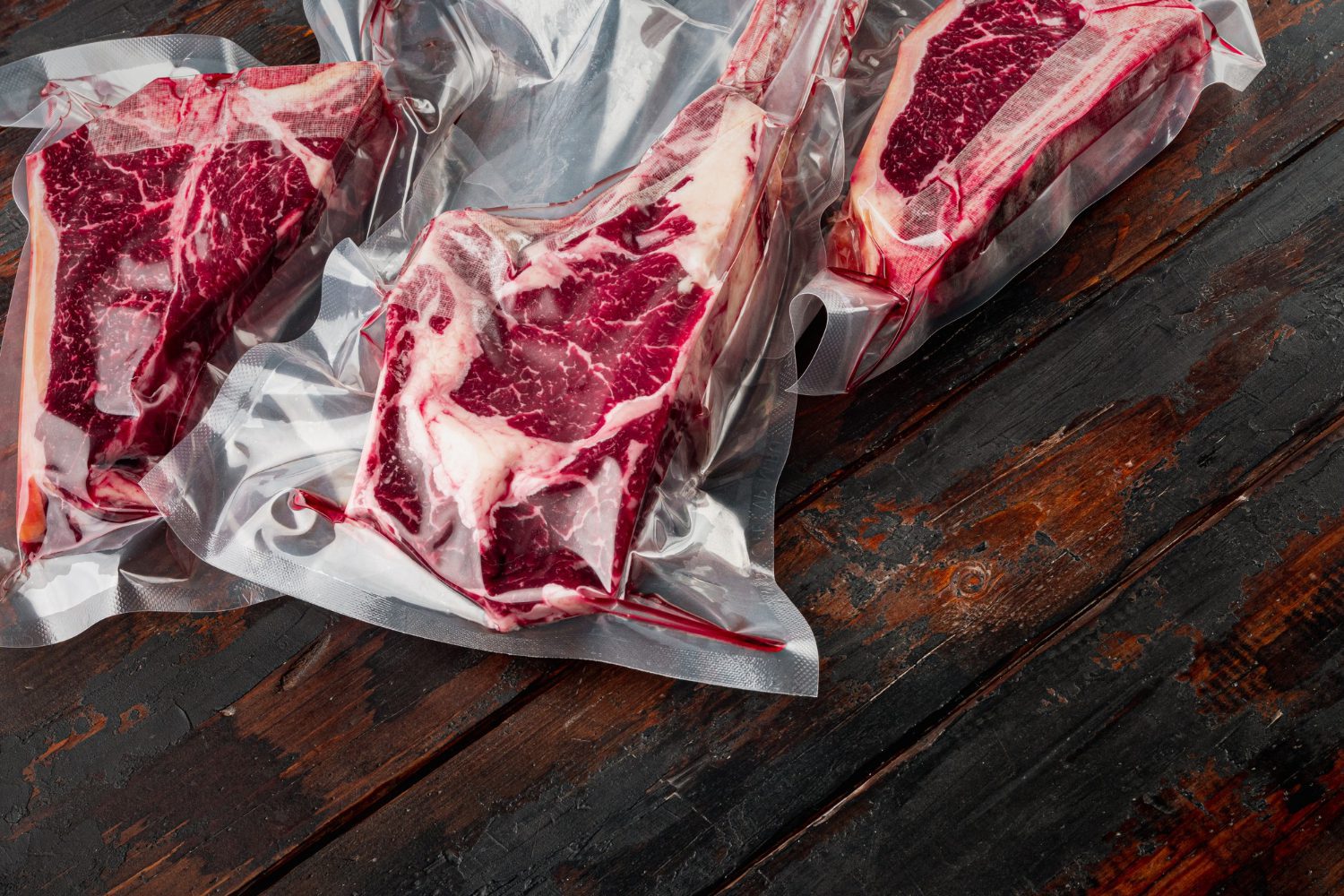 Three raw steaks in vacuum-sealed plastic packaging on a dark wooden surface.