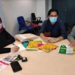 Three people wearing face masks sitting at a table with documents and office supplies.